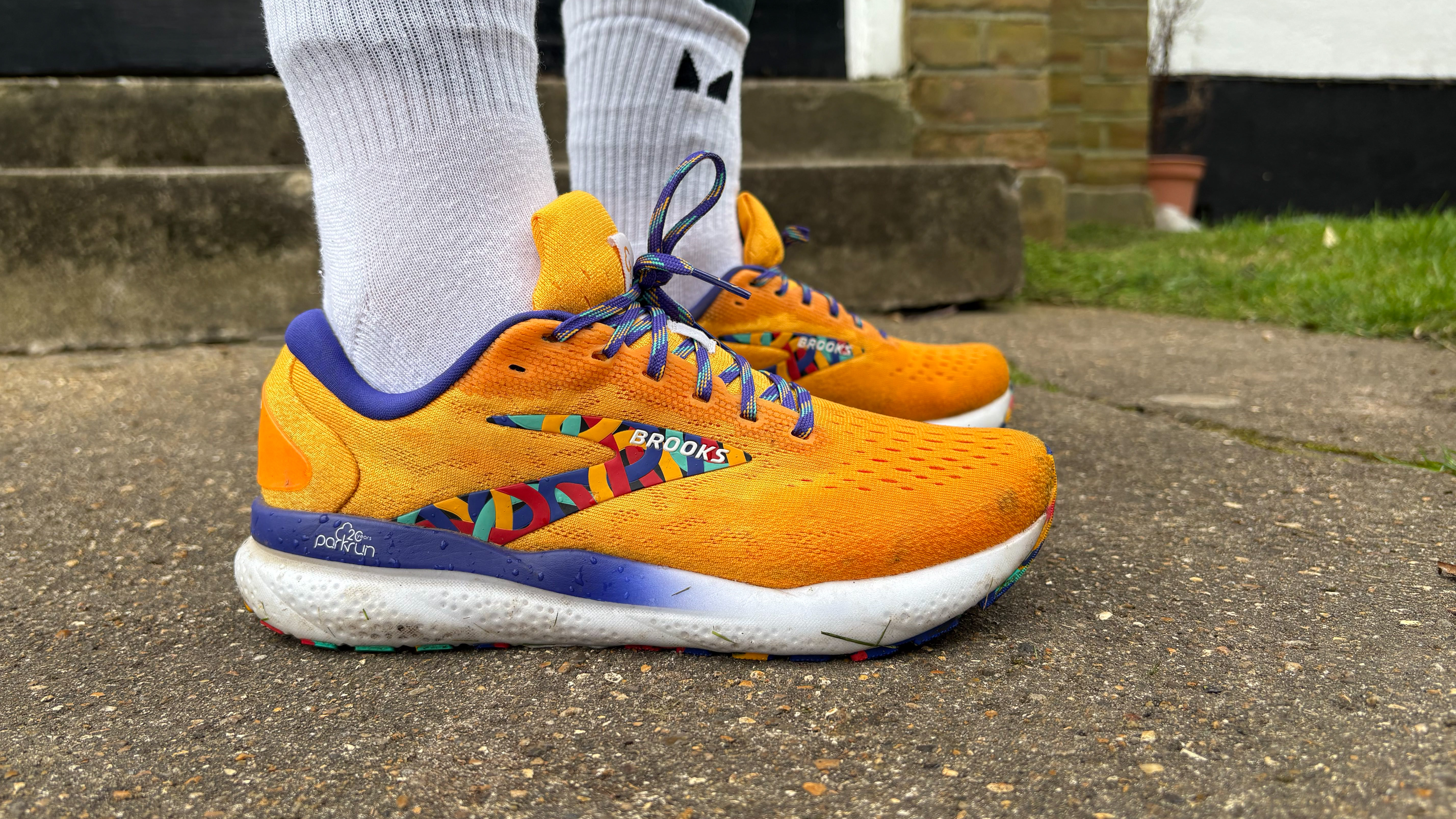 Close up on a pair of running sneakers (the Brooks Ghost 16) being worn by fitness editor Ruth Gaukrodger. The shoes are brightly colored, with contrasting side panels and laces. Ruth wears long socks and behind her we can see steps and short cut grass.