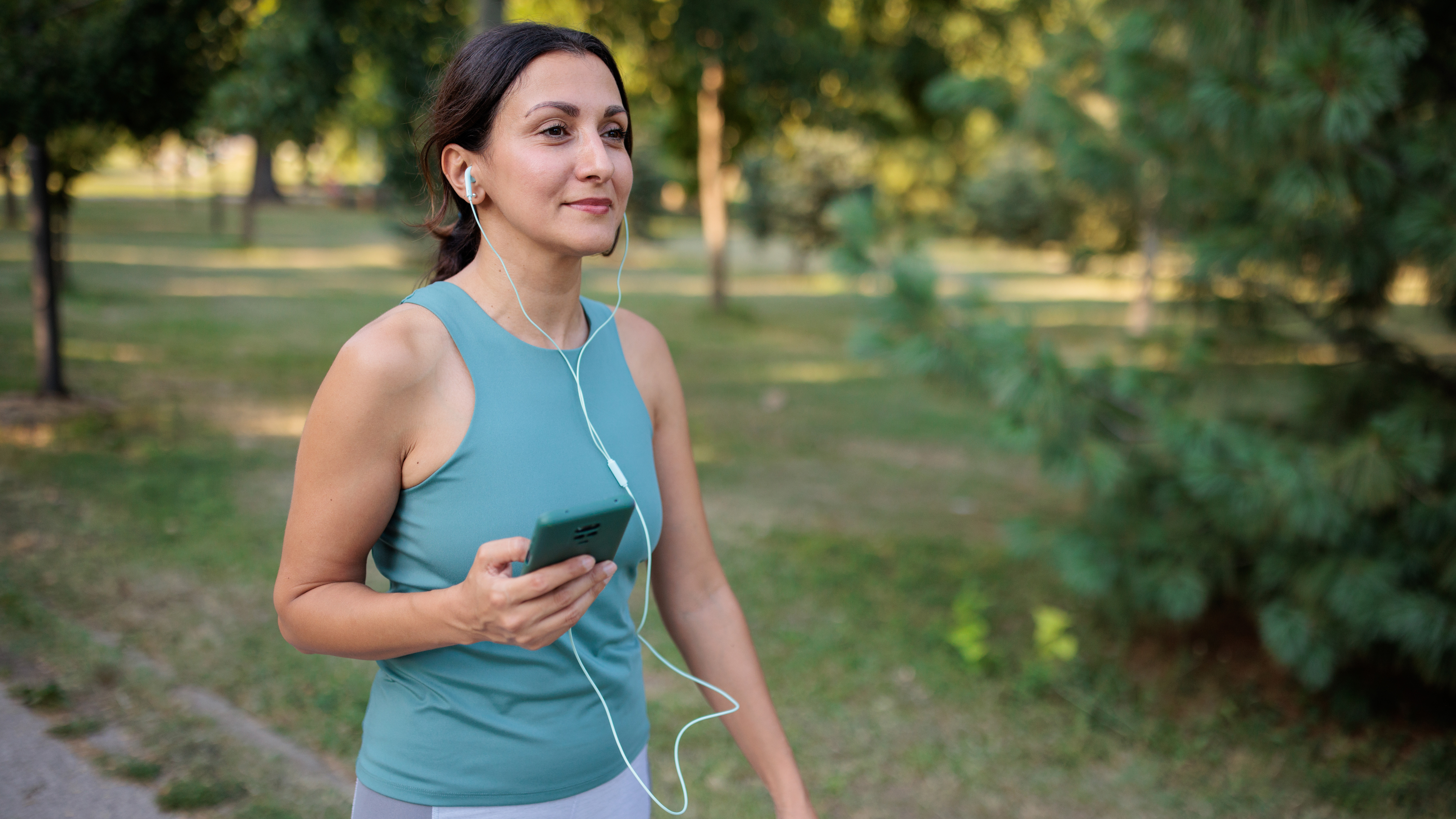 A woman walks outside smiling and wearing headphones, which are plugged into a phone she holds in her right hand. Behind her we see a sunny park with trees.