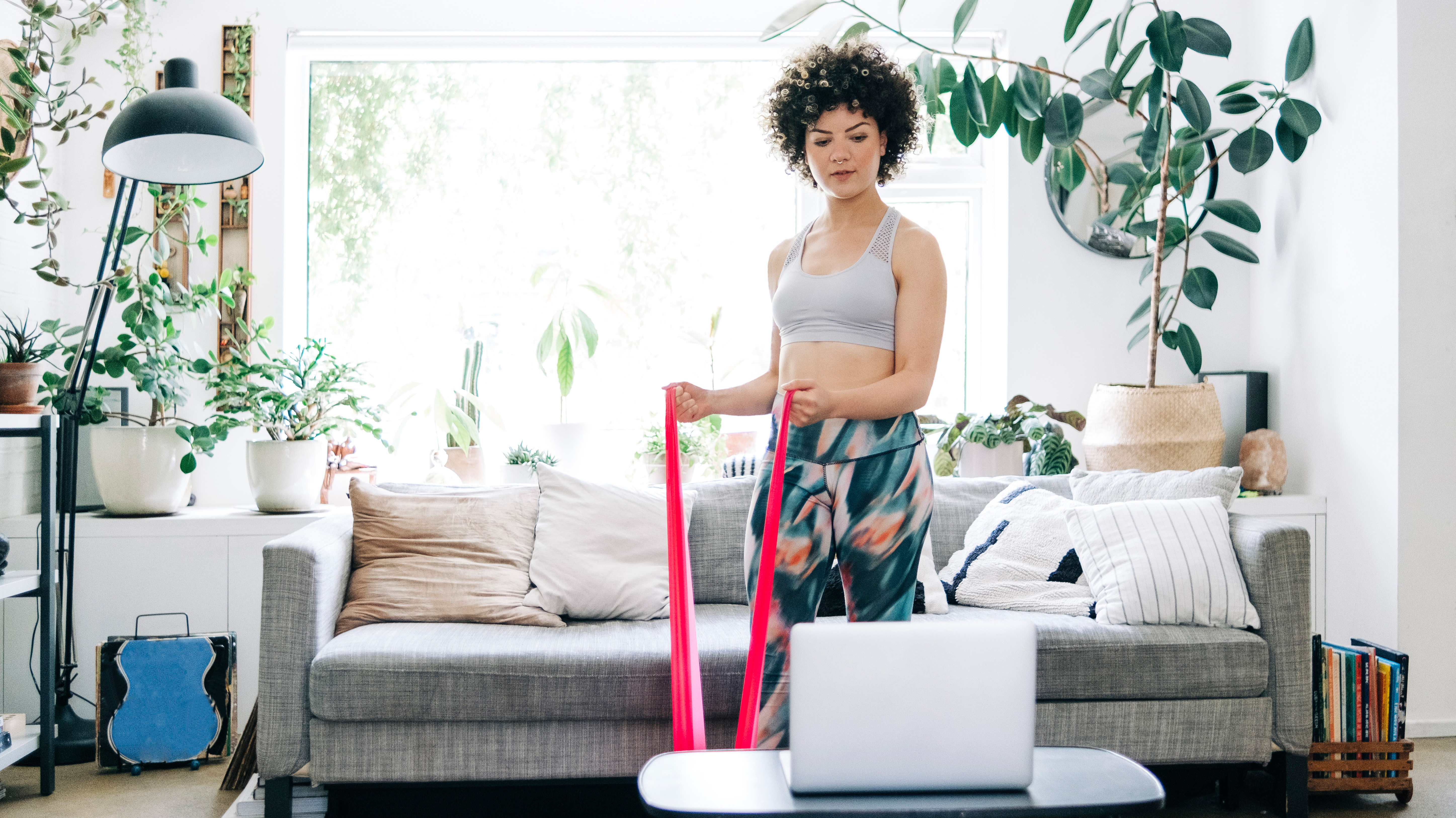 A woman stands in a plant-filled living room in workout clothes, looking at a laptop on a coffee table and holding a resistance band in her hands. The ends of the band is in her hands, while the middle is taught on the floor underneath her foot. Behind her we see a couch and cushions.