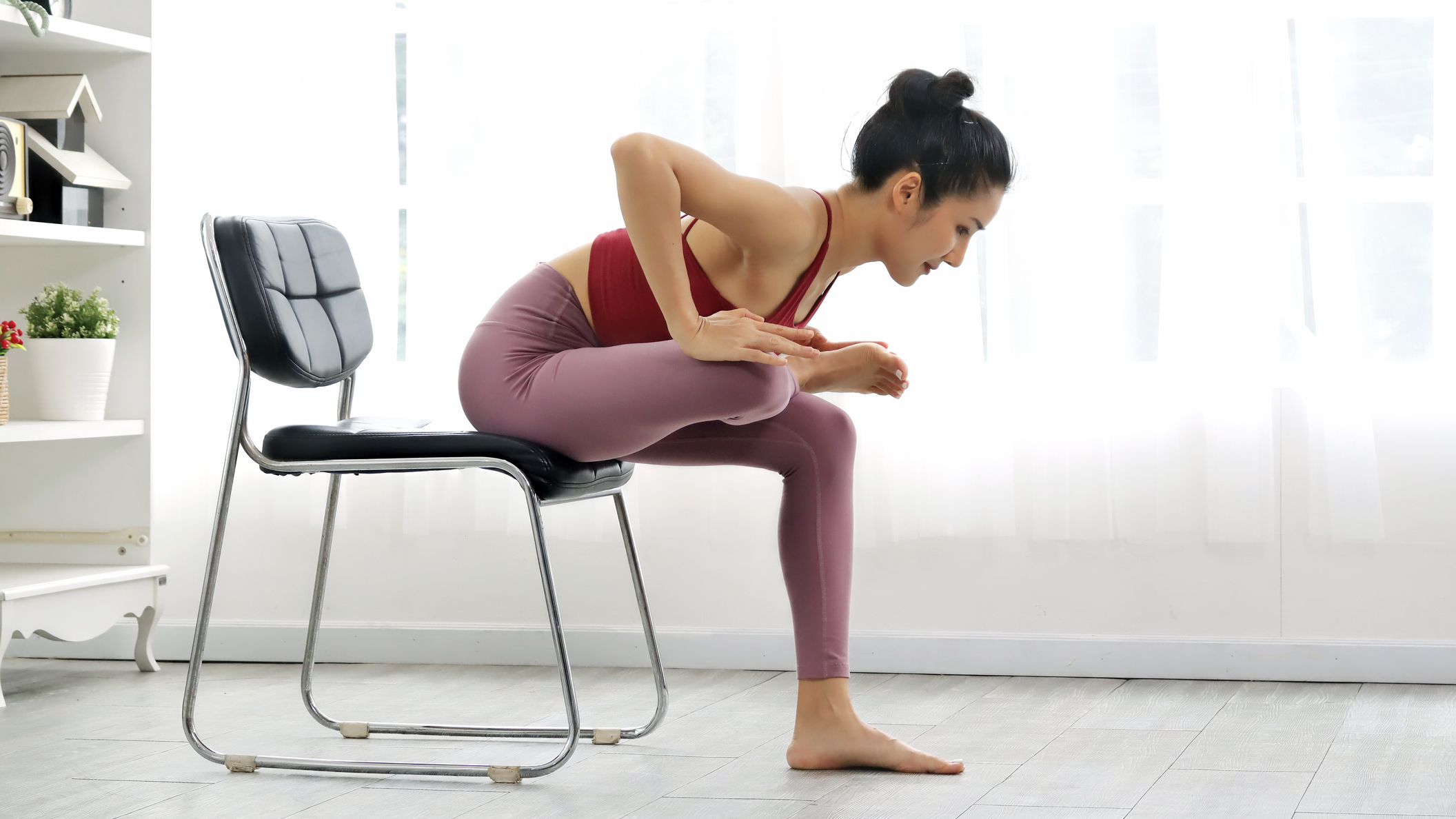Woman in sportswear performs glute stretch while sitting in a chair
