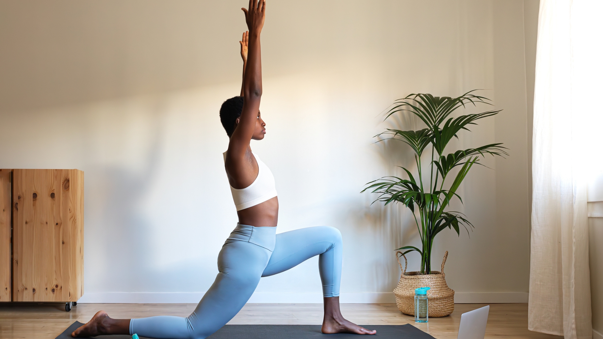 woman performing a low lunge with arms extended overhead, wearing blue leggings and white croptop in a living room setting with a plant to one side