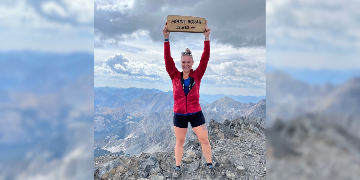 Woman in sportswear stands on top of mountain holding sign that reads "MOUNT BORAH 12,662 ft" triumphantly above her head