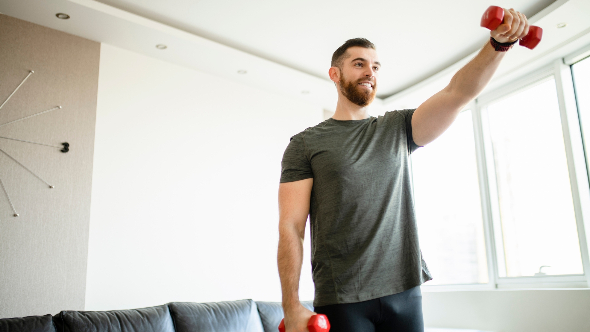 man wearing a grey tshirt raising one arm in front holding a small red dumbbell. there's a plain wall and a window behind him in a living room setting.