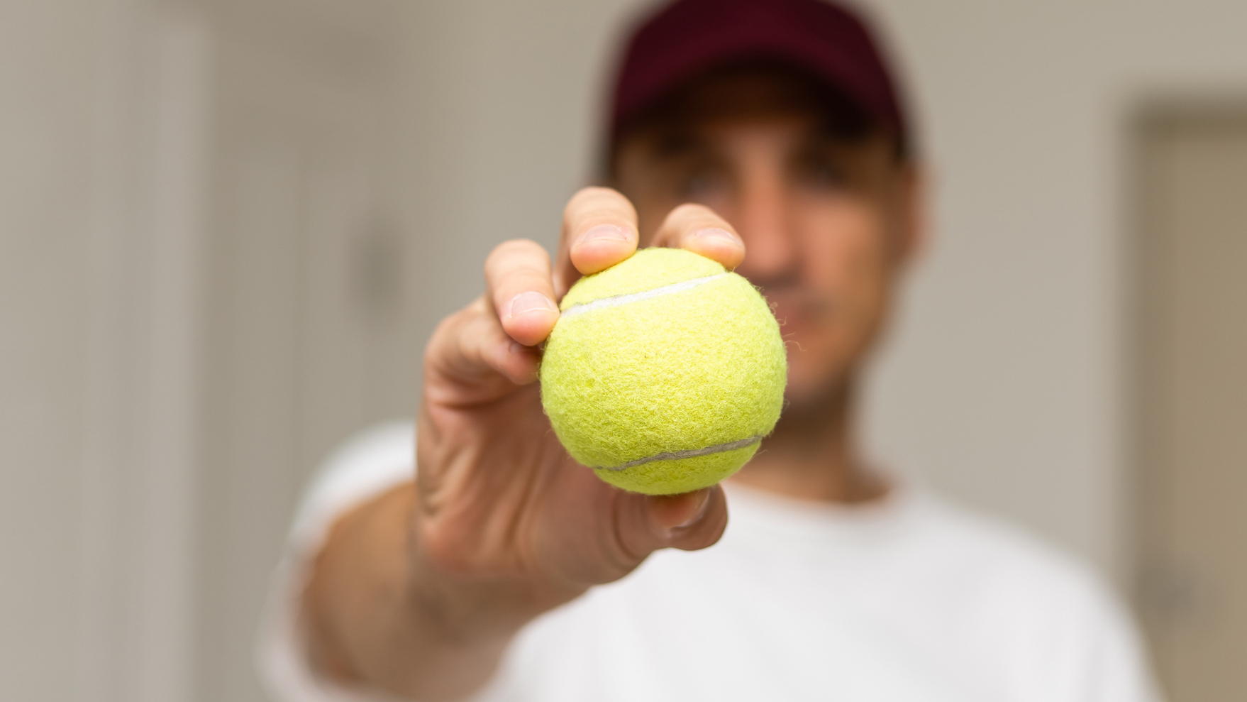 Man holding up tennis ball to viewer. He is inside and wears a white T-shirt and red cap