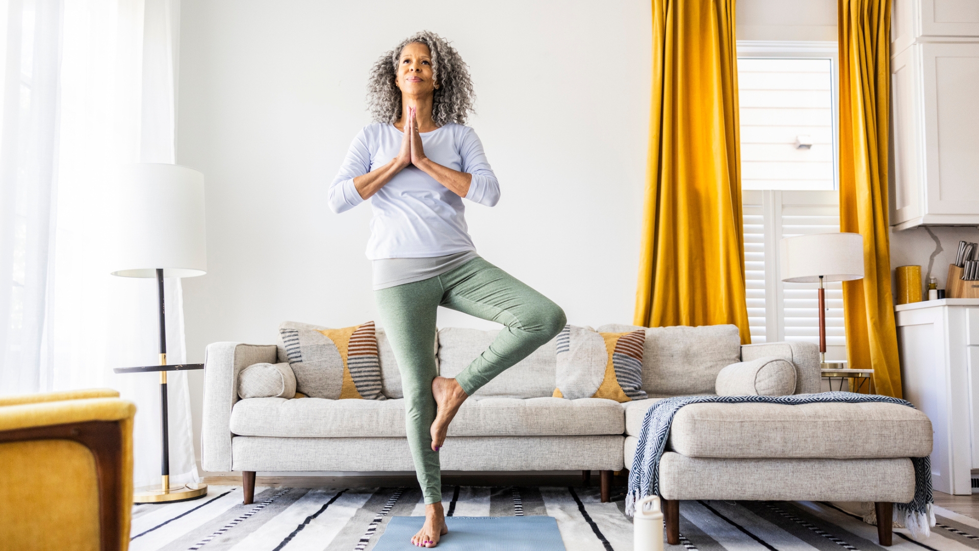woman holding a tree yoga pose in a living room setting with a grey L-shape sofa behind her and a window with long yellow curtains.