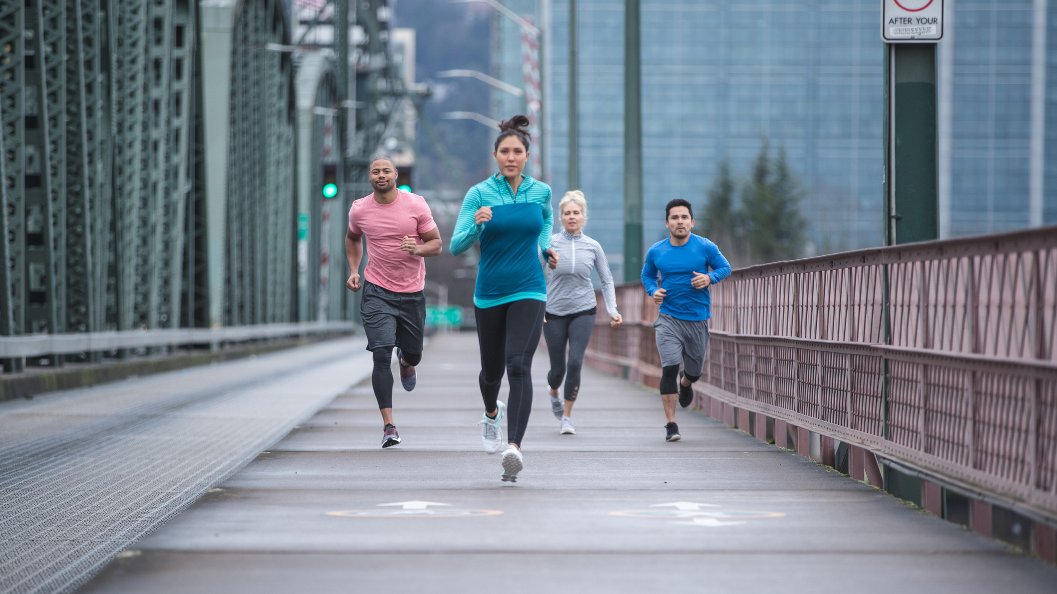 Four runners run across bridge and towards camera, there are tall office buildings in the background