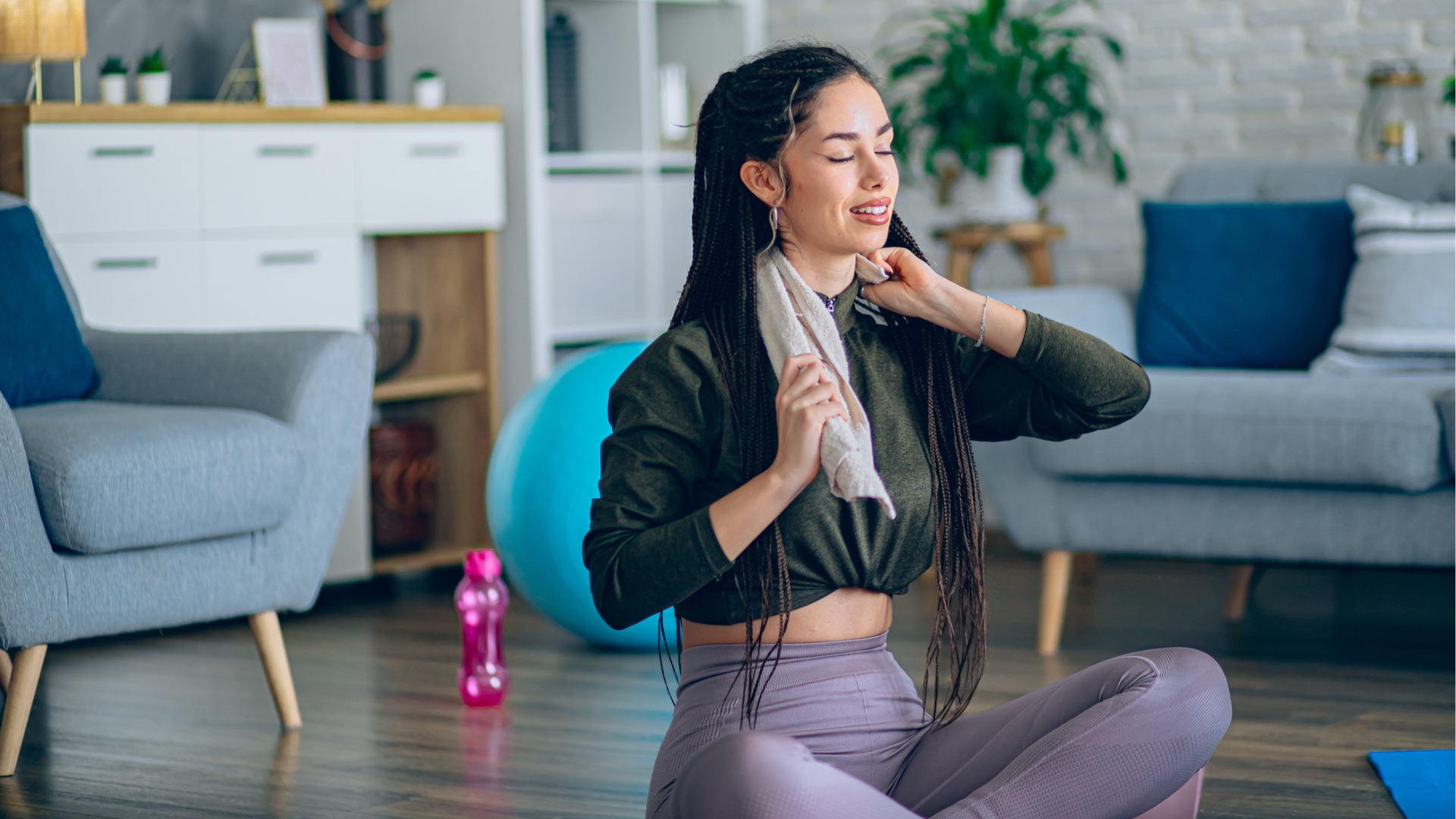 woman sat cross legged on the floor in a living room holding a towel around her neck, smiling with her eyes closed