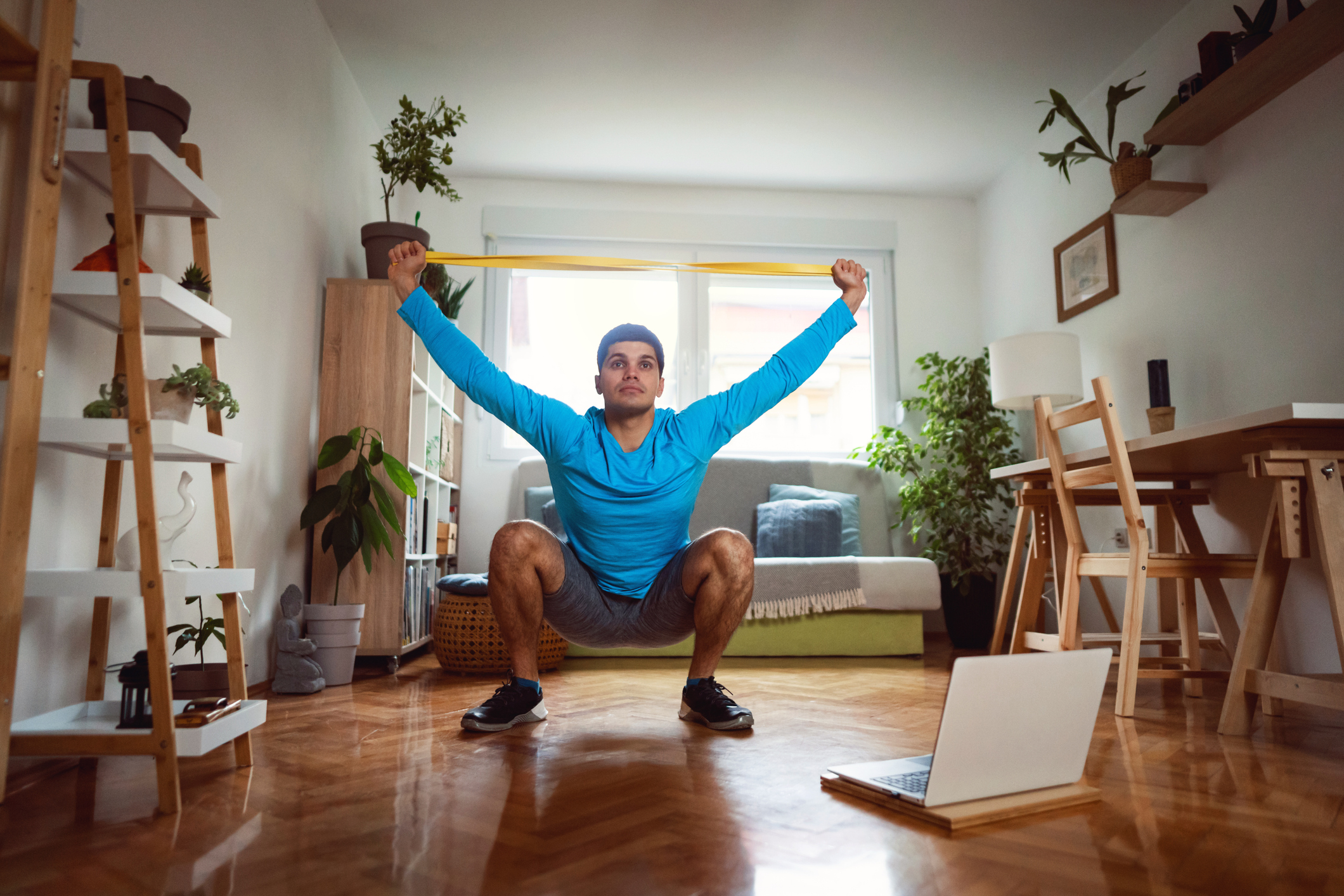 Man in sportswear in living room sits in a deep squat holding a resistance band in both hands above his head.