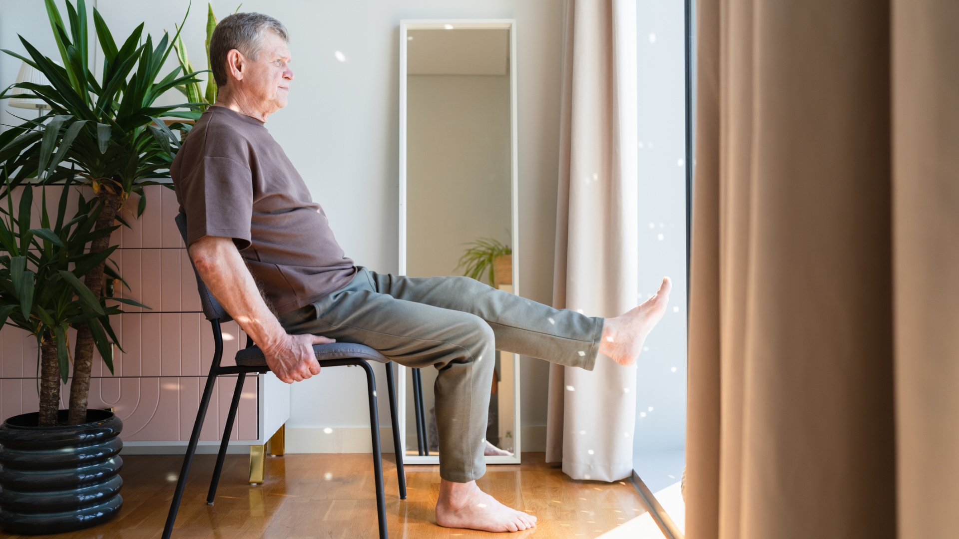 man on a chair and wooden floor, sat sideways to the camera, looking out into a window with a plant behind him. he has one leg raised