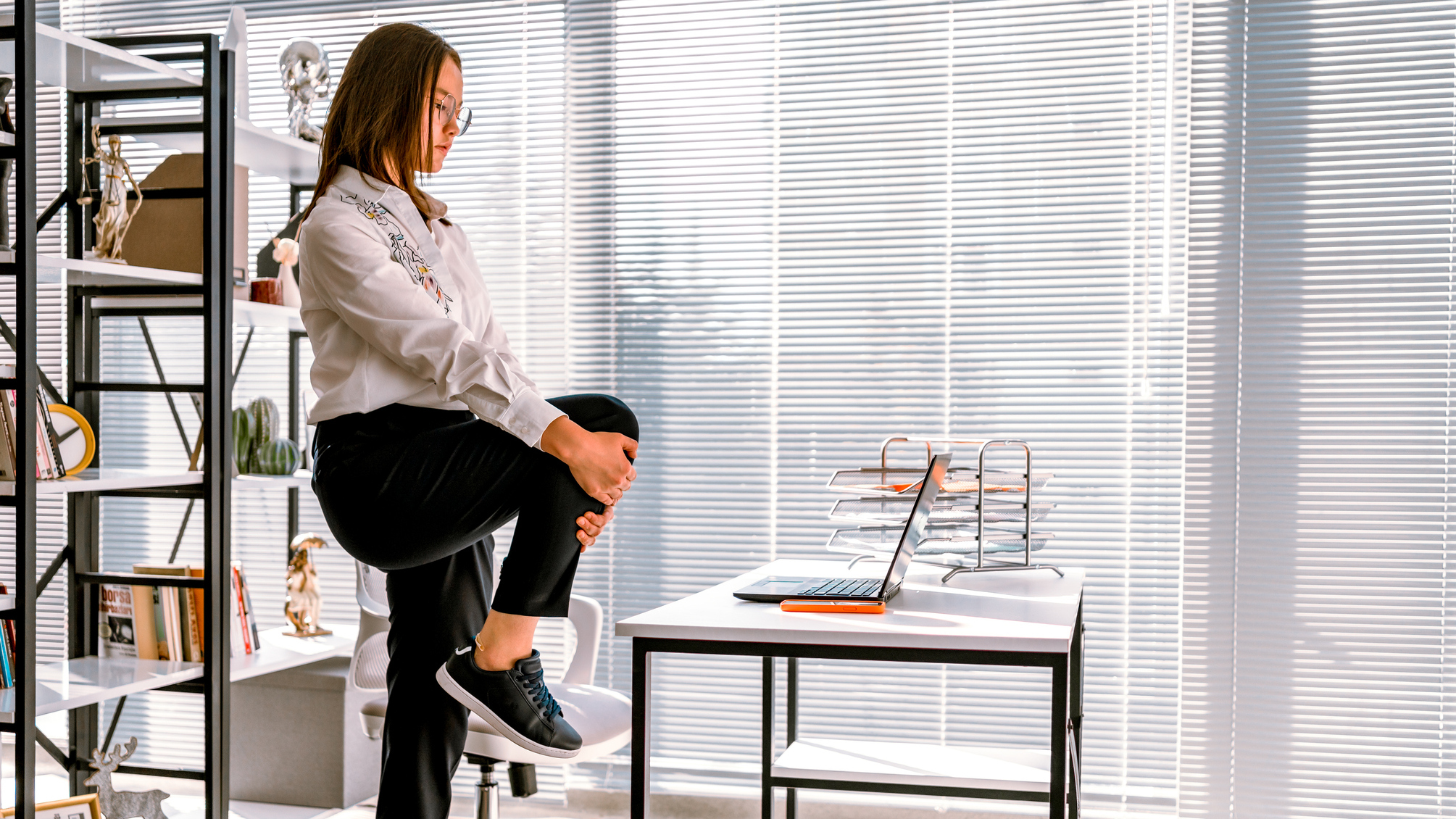 Woman stands on one leg stretching in front of laptop on desk