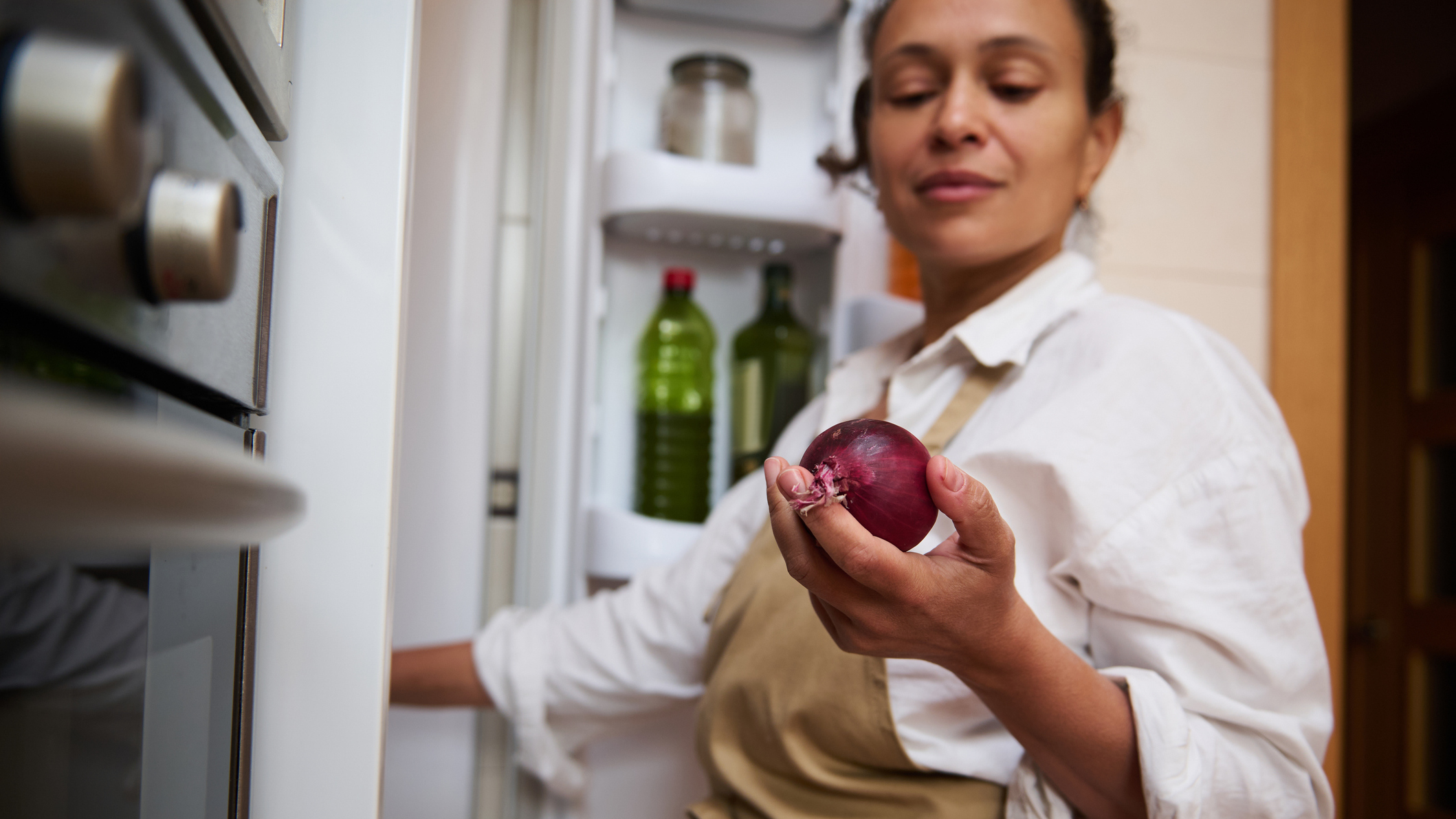 Woman wearing a white shirt and a brown apron stands in front of open fridge holding a red onion