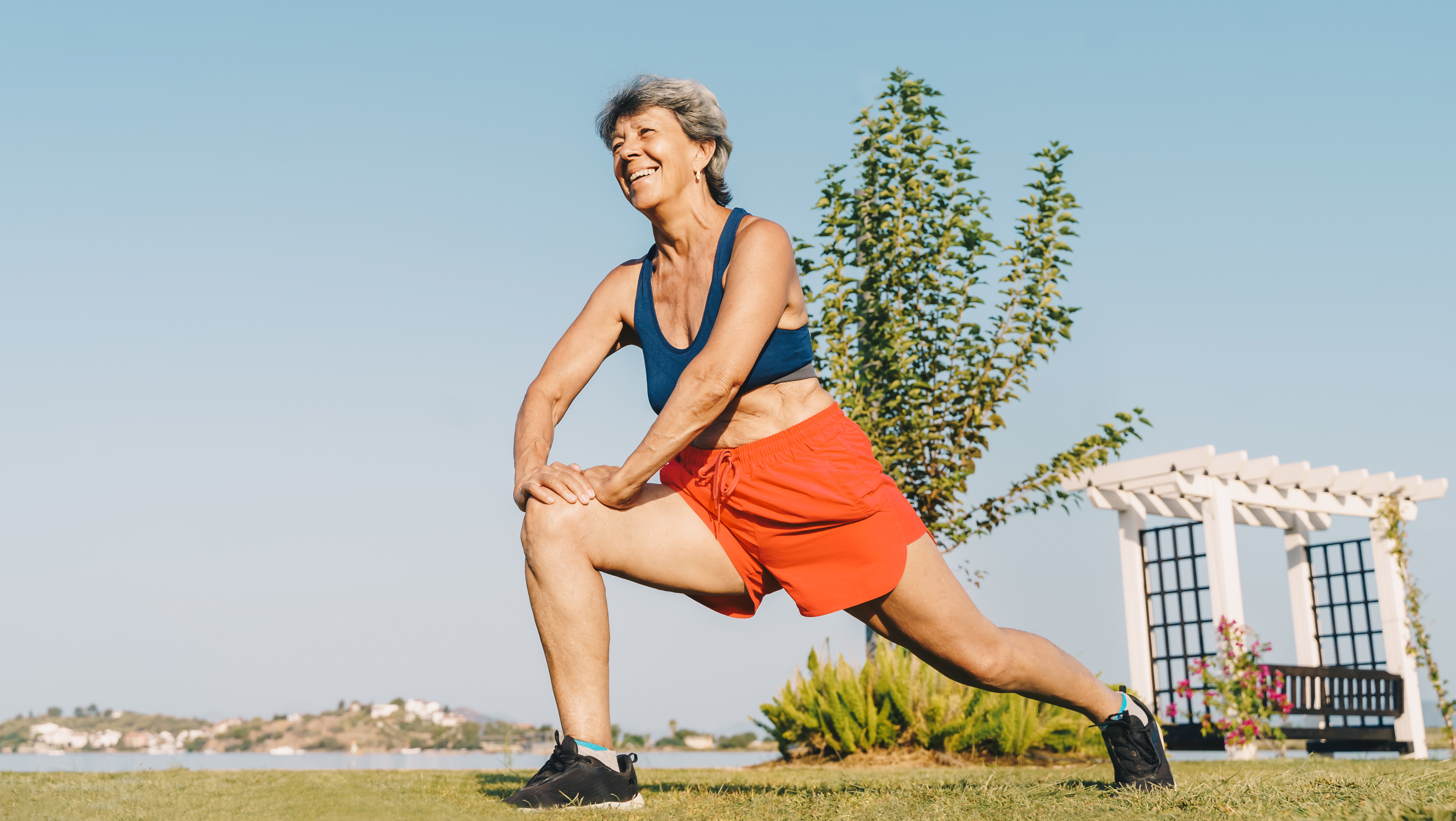 A woman performs a lunge outside in shorts and a sports bra. She is smiling and the sky is clear. Behind her we see a body of water and some distant buildings.