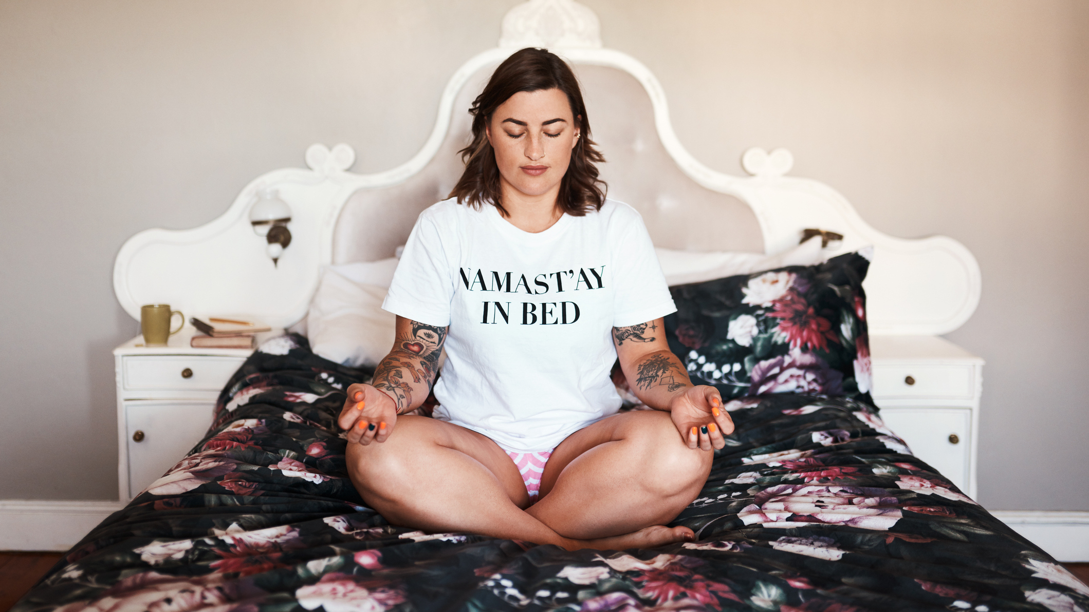 Woman sits cross-legged on a bed spead wearing a white T-shirt that reads NAMAST’AY IN BED