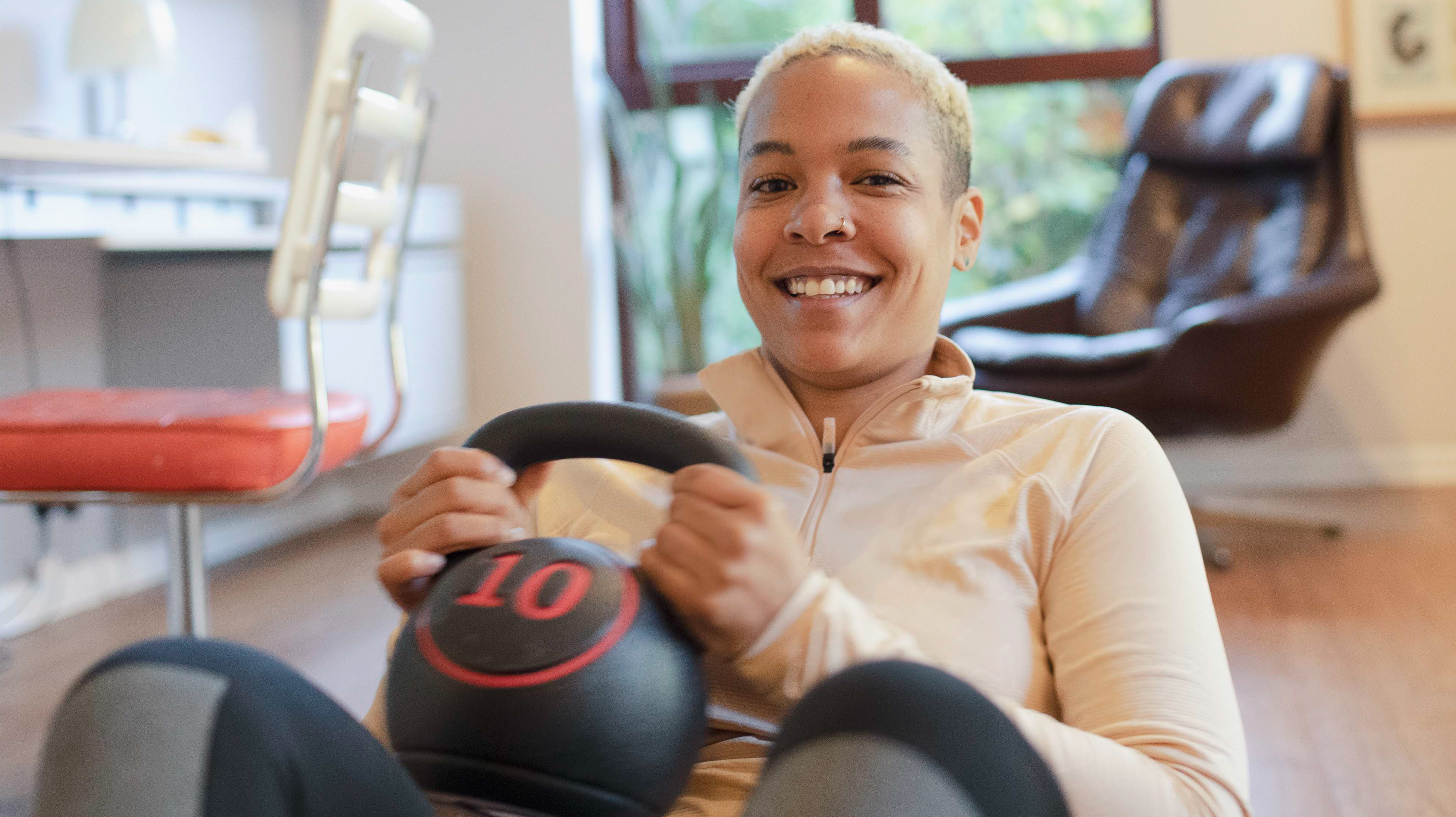 A smiling woman works out at home with a kettlebell. She is sat on the floor with the kettlebells in her hands as she performs a sit-up. Behind her are two desk chairs.